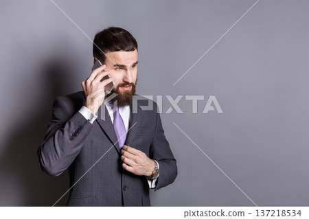 Concentrated bearded businessman talking on phone. Young serious man in suit having important conversation, gray studio background, copy space. Communication concept Concentrated bearded businessman talking on phone. Young serious man in suit having important conversation, gray studio background, copy space. Communication concept 137218534
