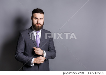 Handsome young bearded man in suit adjusting sleeves. Confident stylish businessman ready for meeting, copy space, gray studio background Handsome young bearded man in suit adjusting sleeves. Confident stylish businessman ready for meeting, copy space, gray studio background 137218548
