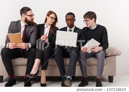 Four individuals are gathered on a couch in an office. Two men and two women are focused on a laptop. One man holds a notebook, while another has a tablet. They are discussing ideas. Four individuals are gathered on a couch in an office. Two men and two women are focused on a laptop. One man holds a notebook, while another has a tablet. They are discussing ideas. 137218623