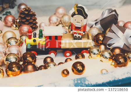 Christmas gifts background. Vintage wooden toy train and nutcracker on snow with golden shiny baubles and small present boxes. Magical atmosphere of new year eve, closeup Christmas gifts background. Vintage wooden toy train and nutcracker on snow with golden shiny baubles and small present boxes. Magical atmosphere of new year eve, closeup 137218650