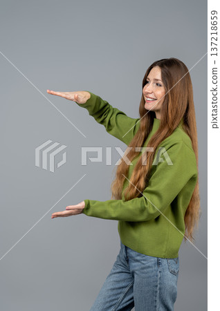 Woman in green clothes showing something with hands on a grey background Woman in green clothes showing something with hands on a grey background 137218659
