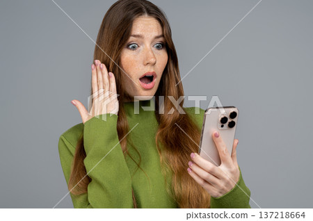 Young woman reading a message on her phone and looking shocked Young woman reading a message on her phone and looking shocked 137218664
