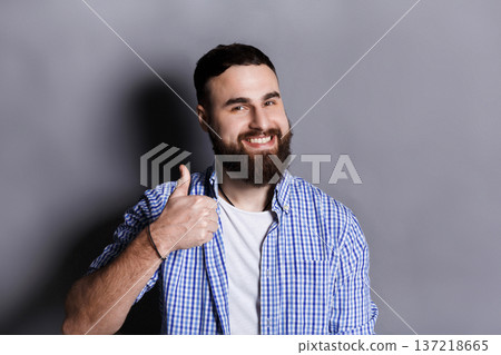 Young hipster man with thumb up portrait. Cheerful guy gesturing that all is great, gray studio background Young hipster man with thumb up portrait. Cheerful guy gesturing that all is great, gray studio background 137218665