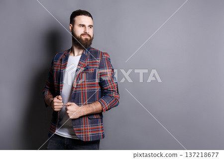 Handsome bearded man looking away, copy space. Portrait of young confident hipster holding his checkered jacket, gray studio background 137218677
