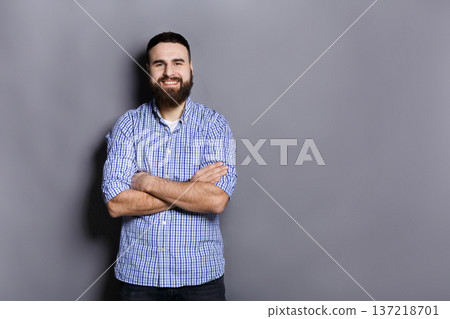 Handsome confident bearded man portrait. Young guy with crossed hands smiling at camera, gray studio background Handsome confident bearded man portrait. Young guy with crossed hands smiling at camera, gray studio background 137218701