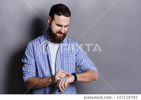 Bearded man clicking on screen of his smart watch. Pleased smiling man enjoying his modern hand clock, gray studio background, copy space Bearded man clicking on screen of his smart watch. Pleased smiling man enjoying his modern hand clock, gray studio background, copy space 137218705