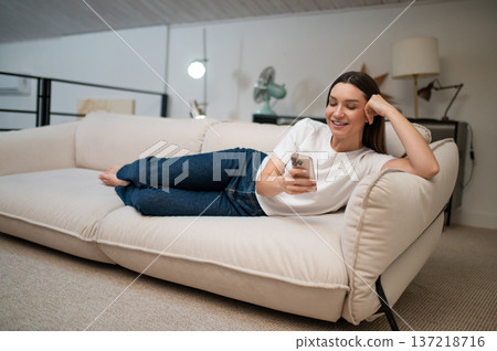 Young woman lying on the sofa with a phone and hand and looking pleased Young woman lying on the sofa with a phone and hand and looking pleased 137218716