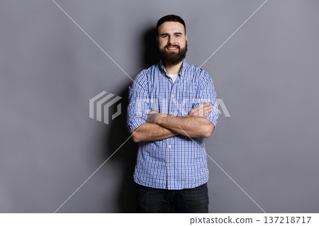 Handsome confident bearded man portrait. Young guy with crossed hands smiling at camera, gray studio background Handsome confident bearded man portrait. Young guy with crossed hands smiling at camera, gray studio background 137218717