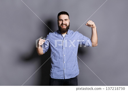 Excited bearded man very glad to success. Young cheerful guy smiling at camera and gesturing with hands, gray studio background 137218730