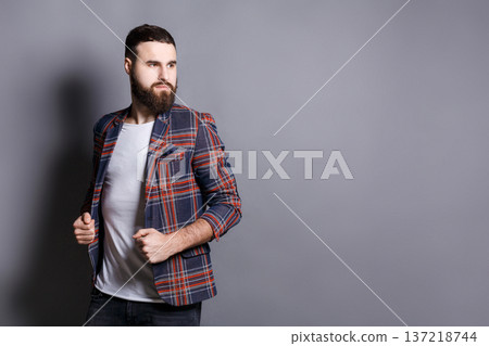 Handsome bearded man looking away, copy space. Portrait of young confident hipster holding his checkered jacket, gray studio background 137218744