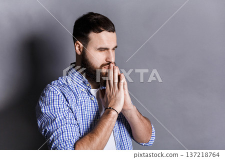 Portrait of hopeful bearded man praying. Young guy holding hands clasped near face, looking away, gray studio background 137218764