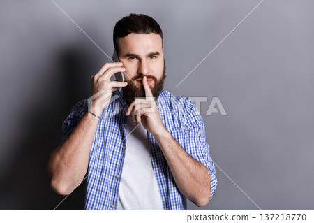 Bearded man talking on phone point be quiet. Young guy having important conversation, showing hush sign with finger on lips, gray studio background, copy space Bearded man talking on phone point be quiet. Young guy having important conversation, showing hush sign with finger on lips, gray studio background, copy space 137218770