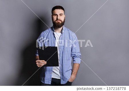 Young serious bearded man standing with folder, preparing for speech or report, gray background, copy space 137218778