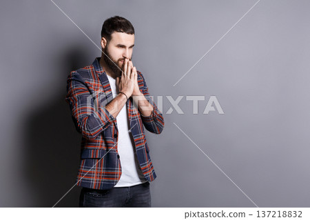 Portrait of hopeful bearded hipster praying. Young guy holding hands clasped near face, looking away, gray studio background, copy space 137218832