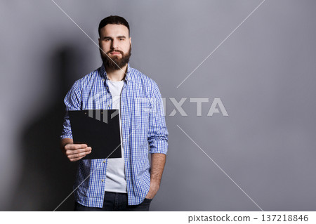 Young serious bearded man standing with folder, preparing for speech or report, gray background, copy space Young serious bearded man standing with folder, preparing for speech or report, gray background, copy space 137218846