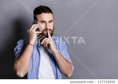Bearded man talking on phone point be quiet. Young guy having important conversation, showing hush sign with finger on lips, gray studio background, copy space Bearded man talking on phone point be quiet. Young guy having important conversation, showing hush sign with finger on lips, gray studio background, copy space 137218856