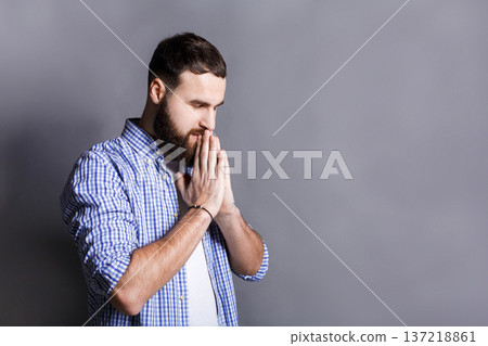 Portrait of hopeful bearded man praying. Young guy holding hands clasped near face, looking away, gray studio background 137218861