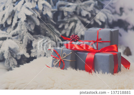 Christmas gifts decorated with red ribbon bows on soft fluffy carpet at blurred snowy fir tree background, closeup, selective focus. Winter holidays concept Christmas gifts decorated with red ribbon bows on soft fluffy carpet at blurred snowy fir tree background, closeup, selective focus. Winter holidays concept 137218881