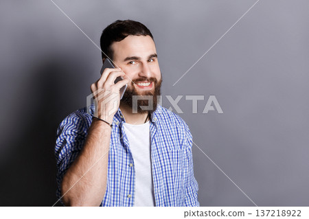 Cheerful bearded man talking on phone. Young smiling guy having nice conversation, gray studio background, copy space. Communication concept 137218922