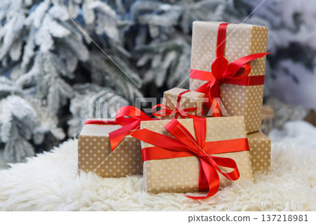 Christmas gifts decorated with red ribbon bows on soft fluffy carpet at blurred snowy fir tree background, closeup, selective focus. Winter holidays concept Christmas gifts decorated with red ribbon bows on soft fluffy carpet at blurred snowy fir tree background, closeup, selective focus. Winter holidays concept 137218981