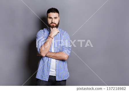 Pensive bearded man posing with hand on chin. Portrait of young confident guy, looking at camera, gray studio background 137218982