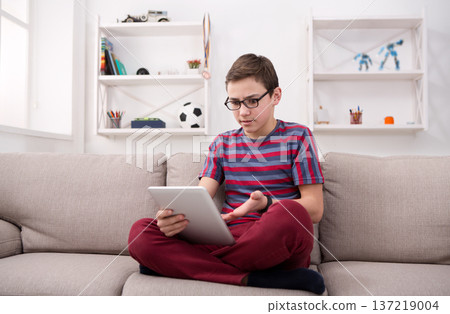 Gadgets and technology concept - teenage boy using digital tablet, watching football on portable device, playing online game while sitting on the couch in living room at home 137219004