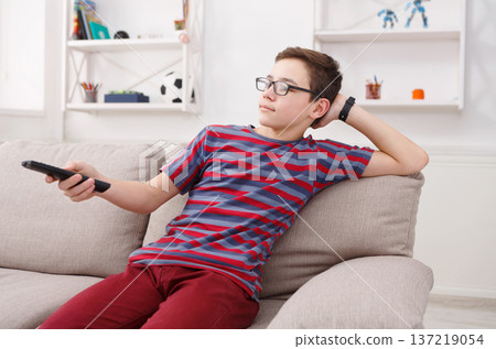 Teenager boy watching television, using TV remote while sitting on couch in living room at home 137219054