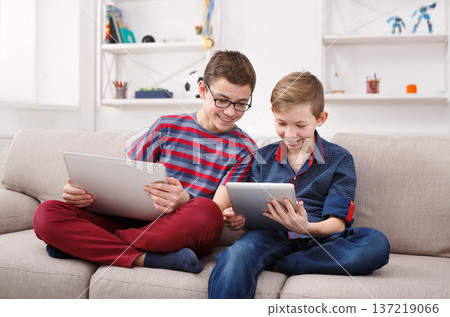 Technology, boy friendship and communication concept - two cute teenagers using tablets, playing games, talking and smiling while sitting on the couch in living room at home 137219066