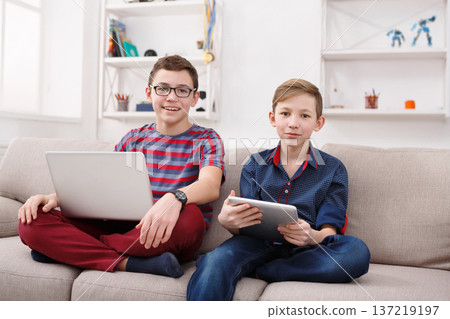 Two kids with gadgets looking at camera. Stylish boys with laptop and digital tablet on sofa at home. Boy friendship and communication concept Two kids with gadgets looking at camera. Stylish boys with laptop and digital tablet on sofa at home. Boy friendship and communication concept 137219197