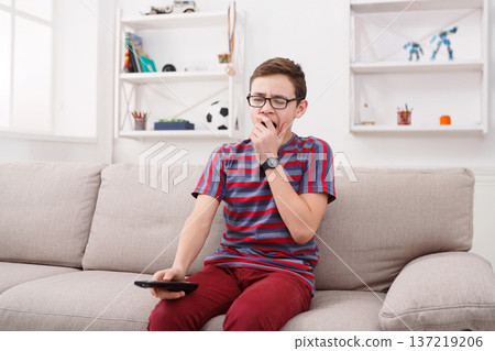 Boring television. Teenager holding TV remote and looking bored while sitting the couch at home in living room Boring television. Teenager holding TV remote and looking bored while sitting the couch at home in living room 137219206