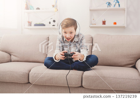 Gaming video games concept, excited boy playing football game with joystick and headphones, enjoying win while sitting on sofa in living room at home 137219239