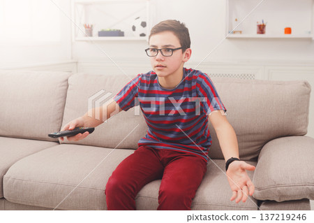 Teenager boy watching television, using remote control to switch channels. Youngster unhappy at what he sees on TV screen. Sitting on couch in living room at home Teenager boy watching television, using remote control to switch channels. Youngster unhappy at what he sees on TV screen. Sitting on couch in living room at home 137219246