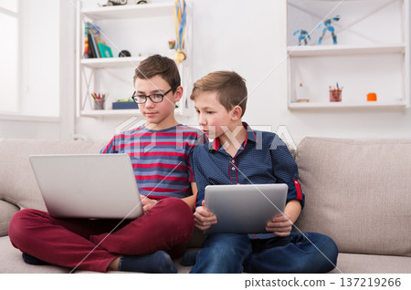 Two male kids with gadgets. Sharing funny content on laptop and digital tablet on sofa at home. Boy friendship and communication concept 137219266