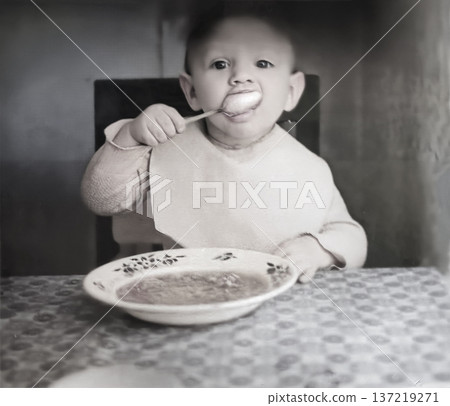 A vintage portrait of a little boy eating from a plate with a spoon. Retro photo from the 1960s. A vintage portrait of a little boy eating from a plate with a spoon. Retro photo from the 1960s. 137219271