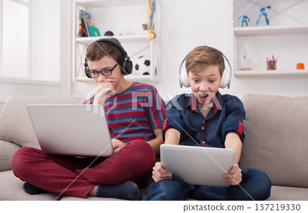 Two male kids with gadgets and headphones. Teenage friends playing games on laptop and digital tablet with excitement at home. Boy friendship and online gaming concept 137219330