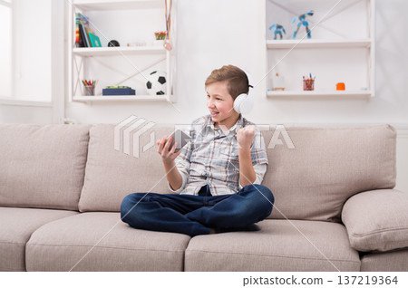 Excited teenage boy with braces playing football game on smartphone and listening to music in headphones, enjoying win while sitting on sofa in living room at home Excited teenage boy with braces playing football game on smartphone and listening to music in headphones, enjoying win while sitting on sofa in living room at home 137219364