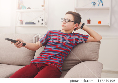 Teenager boy watching television, using TV remote while sitting on couch in living room at home Teenager boy watching television, using TV remote while sitting on couch in living room at home 137219396