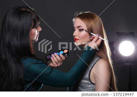 Backstage, fashion shooting. Make-up artist working with beauty model, applying eyeshadow, dark studio background Backstage, fashion shooting. Make-up artist working with beauty model, applying eyeshadow, dark studio background 137219447