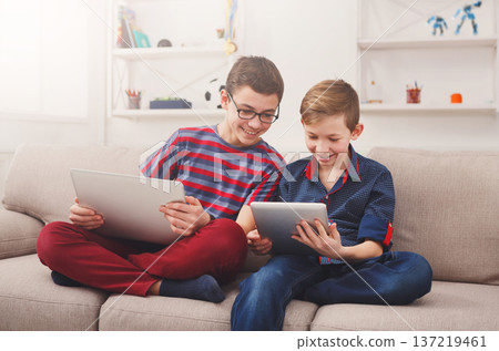 Technology, boy friendship and communication concept - two cute teenagers using tablets, playing games, talking and smiling while sitting on the couch in living room at home 137219461