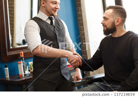 Barber and thankful bearded client handshake. Making modern hairstyle in male barbershop 137219659