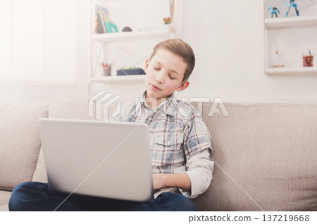 Bored boy with computer on sofa at home. Child trying to find interesting content on the internet, typing on keyboard Bored boy with computer on sofa at home. Child trying to find interesting content on the internet, typing on keyboard 137219668