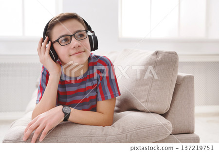 Leisure activity, teenager enjoy music - portrait of dreamy kid in headphones while lying on sofa indoors. Relax time - teenage boy listen to music on couch at home 137219751