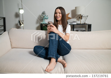 Young woman sitting on the sofa with a phone in hands Young woman sitting on the sofa with a phone in hands 137219781