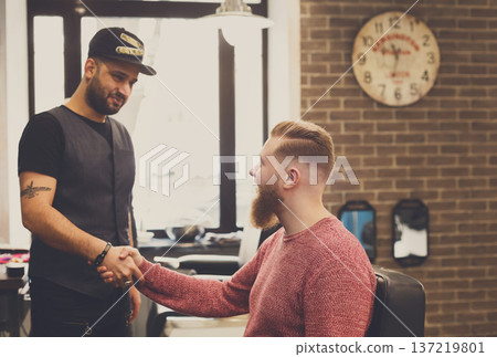 Barber and thankful bearded client handshake. Making modern hairstyle in male barbershop Barber and thankful bearded client handshake. Making modern hairstyle in male barbershop 137219801