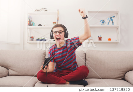 Gaming video games concept - excited teenage boy playing football game with joystick and headphones, enjoying win while sitting on sofa in living room at home 137219830