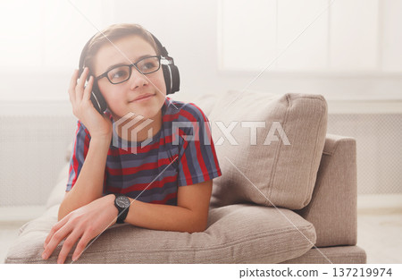 Teenager enjoy music - portrait of dreamy kid in headphones while lying on sofa indoors. Relax time - teenage boy listen to music on couch at home Teenager enjoy music - portrait of dreamy kid in headphones while lying on sofa indoors. Relax time - teenage boy listen to music on couch at home 137219974