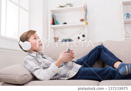 Child in brackets enjoying music in headphones from his smartphone. Boy in casual lying on comfortable sofa, relaxing at home Child in brackets enjoying music in headphones from his smartphone. Boy in casual lying on comfortable sofa, relaxing at home 137219975