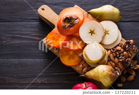 Healthy food - wood board with cut organic raw fruits on wooden background. Top view of sliced ingredients for fruit salad or snack - apple, pear, nuts, persimmon. Flat lay, copy space Healthy food - wood board with cut organic raw fruits on wooden background. Top view of sliced ingredients for fruit salad or snack - apple, pear, nuts, persimmon. Flat lay, copy space 137220211