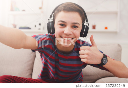 Teenager taking thumb up selfie in headphones on phone while listen to music. Shot of teenage kid gesturing with thumb up while sitting on couch at home 137220249