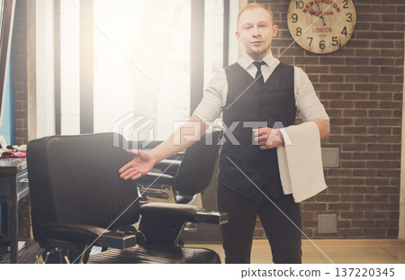 Young elegant barber pointing on a chair, looking at camera and smiling. Male hairstylist inviting customer to have a seat on the chair at barbershop Young elegant barber pointing on a chair, looking at camera and smiling. Male hairstylist inviting customer to have a seat on the chair at barbershop 137220345